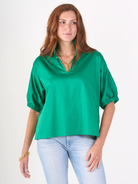 A woman with long red hair and freckles wears the bright green Poppy Top in airy cotton poplin with a V-neck, paired with light blue jeans and gold jewelry, standing against a plain white background.