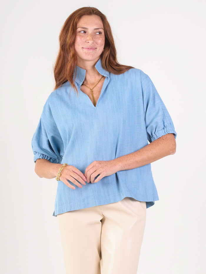 A woman with long red hair wears the Poppy Top in light blue chambray and beige pants. Smiling slightly, she looks off to the side against a plain white background.