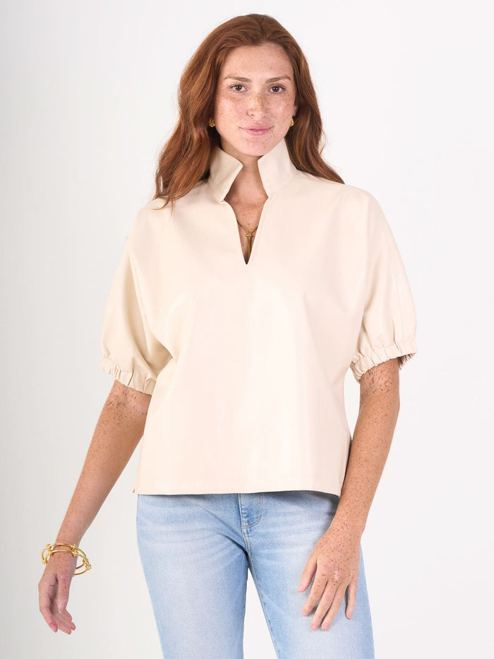 A woman with long red hair smiles gently, standing against a white background in the Emily McCarthy Poppy Top—a light beige, short-sleeved collared blouse with a front slit—paired with light blue jeans.