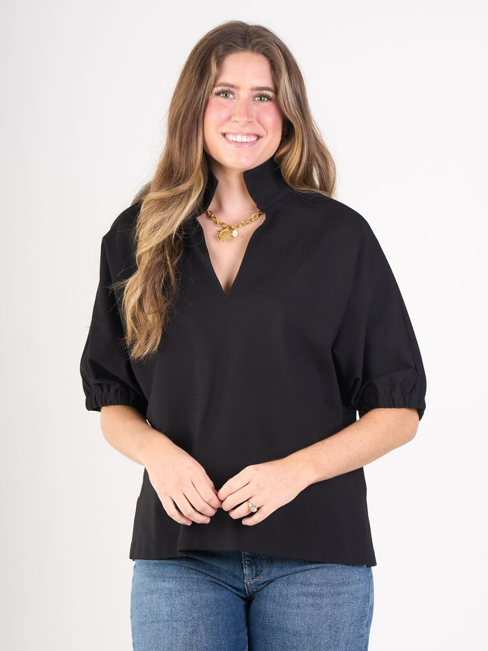 A woman with long brown hair wears the Poppy Top, featuring short sleeves and a high collar, paired with blue jeans and a gold necklace. She smiles while standing against a plain white background.