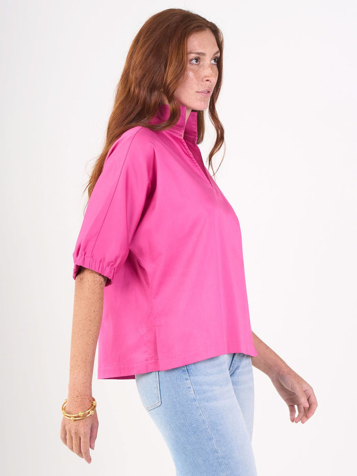 A woman with long red hair stands in profile against a white background, wearing a bright pink Poppy Top blouse, light blue jeans, and gold bracelets.