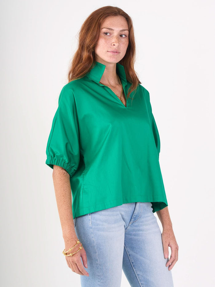 A woman with long red hair wears the Poppy Top in bright green cotton poplin and light blue jeans, standing against a plain white background with a neutral expression, looking slightly to the side.