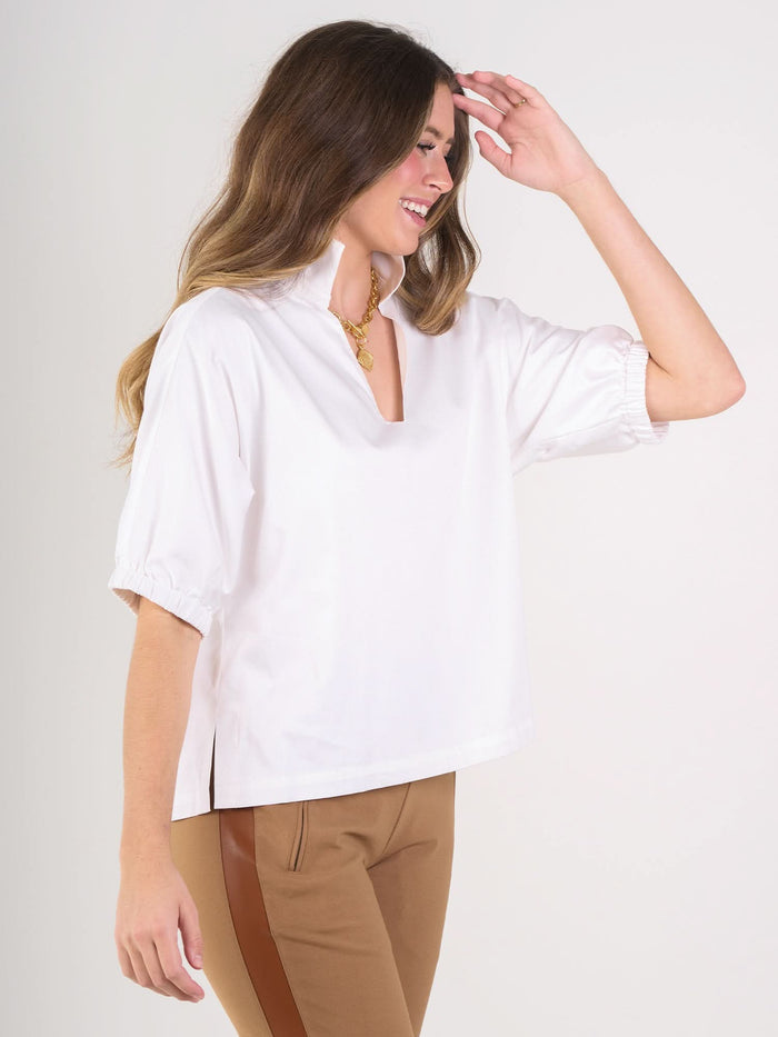 A woman with long brown hair, smiling, wears the white Poppy Top with elbow-length sleeves and tan side-striped pants. She touches her head as she stands against a plain light background.