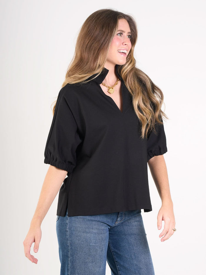 A woman with long brown hair wears the Poppy Top and blue jeans, smiling and standing slightly turned against a plain white background.
