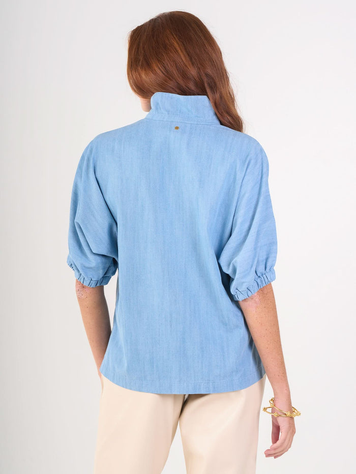 A person with long red hair is shown from the back, wearing the light blue Poppy Top with a high collar and elbow-length puff sleeves, paired with beige pants against a plain white background.