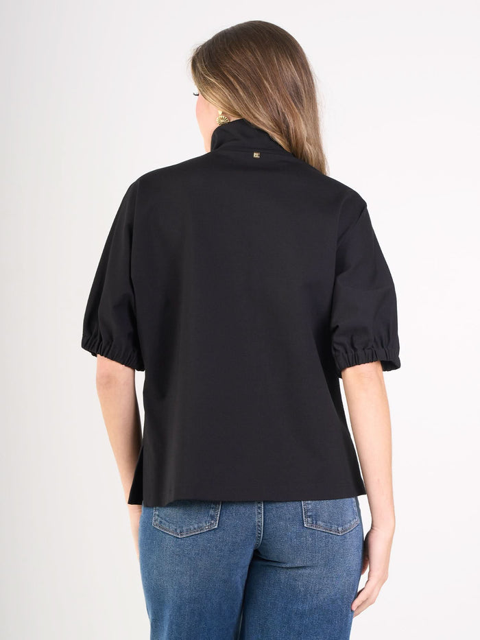 A woman with long brown hair, dressed in a black Poppy Top with puff sleeves and blue jeans, is seen from the back against a plain white background.