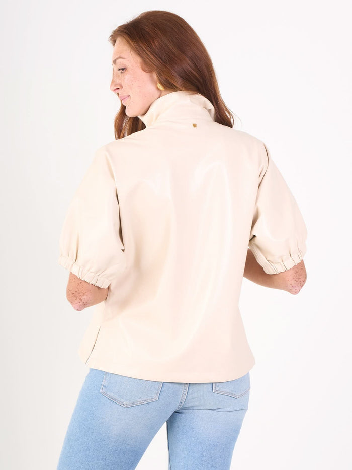 A woman with long auburn hair wears a Poppy Top in light beige and light blue jeans, facing away and looking over her shoulder against a plain white background.