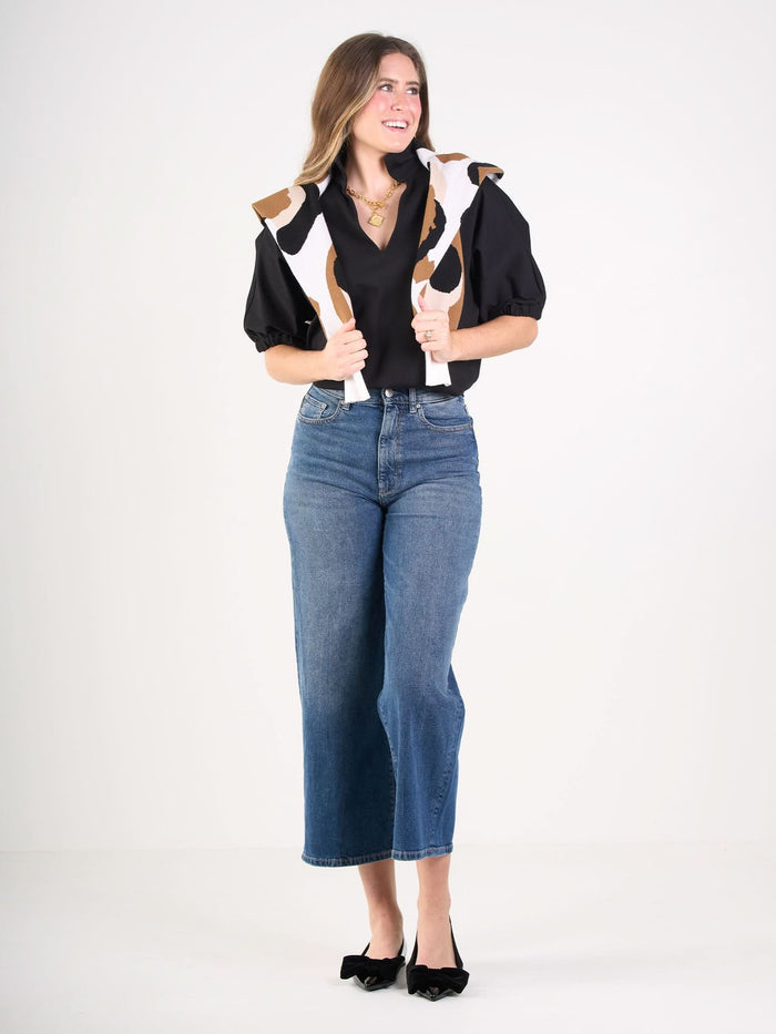 A woman smiles in the Poppy Top, blue high-waisted jeans, black bow shoes, and a brown and white patterned sweater draped over her shoulders, standing against a plain white background.