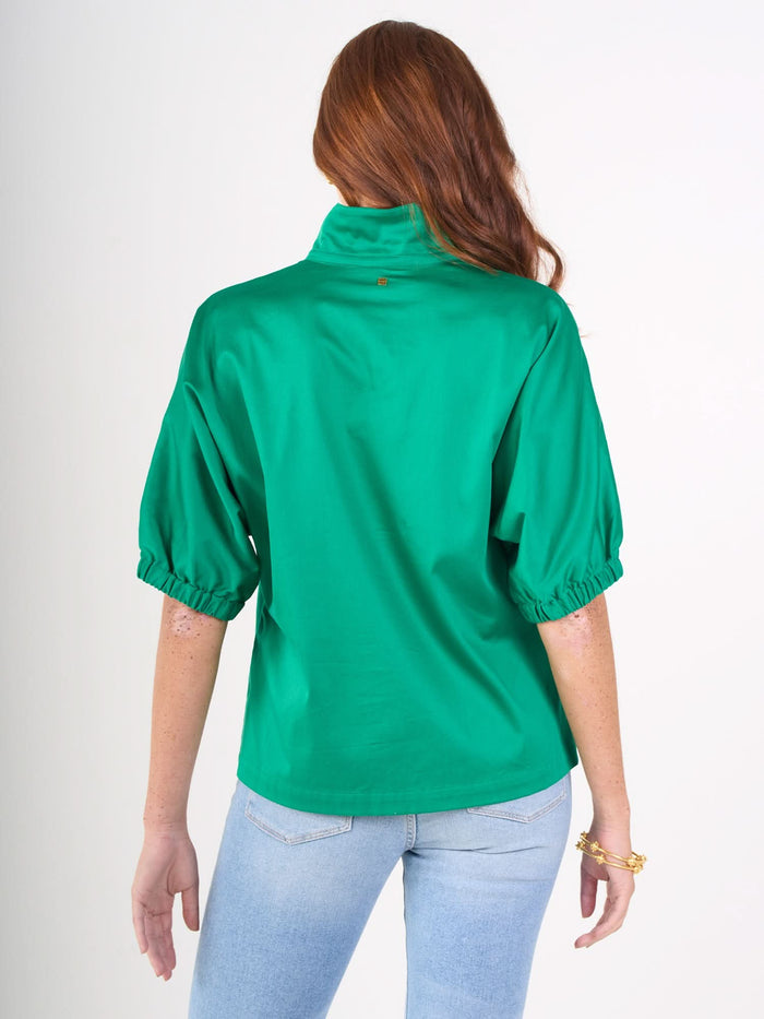 A woman with long, wavy red hair is seen from behind wearing a bright green Poppy Top and light blue jeans, accessorized with a gold bracelet on her left wrist. The background is plain white.