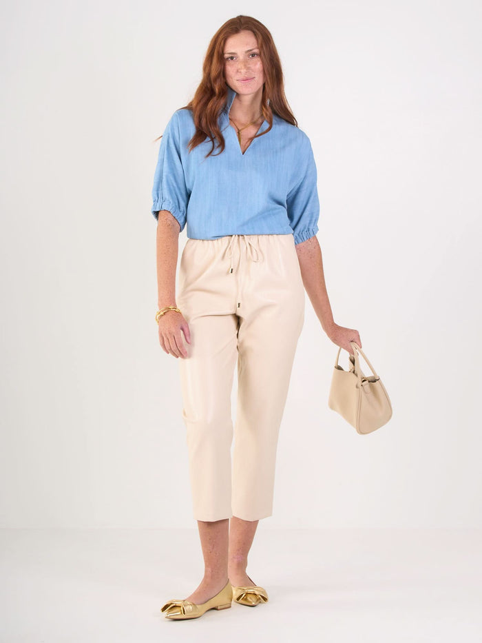 A woman with long red hair wears a Poppy Top in light blue, beige cropped pants, gold flats, and holds a small beige handbag while standing against a plain white background.