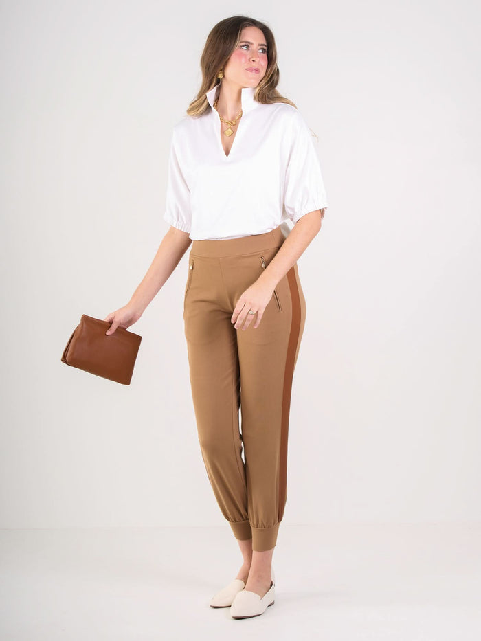 A woman stands against a plain background, wearing the Poppy Top—a loose-fitting white cotton poplin blouse—paired with tan high-waisted pants, cream loafers, a brown clutch, loose hair, and a gold necklace.