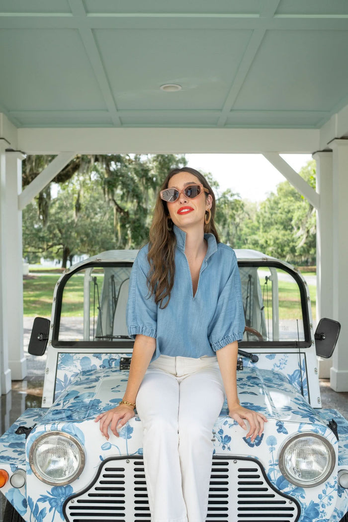 A woman in sunglasses and white pants sits on a vintage car with a blue floral design, wearing the Poppy Top by Emily McCarthy under a covered outdoor area surrounded by trees and greenery.