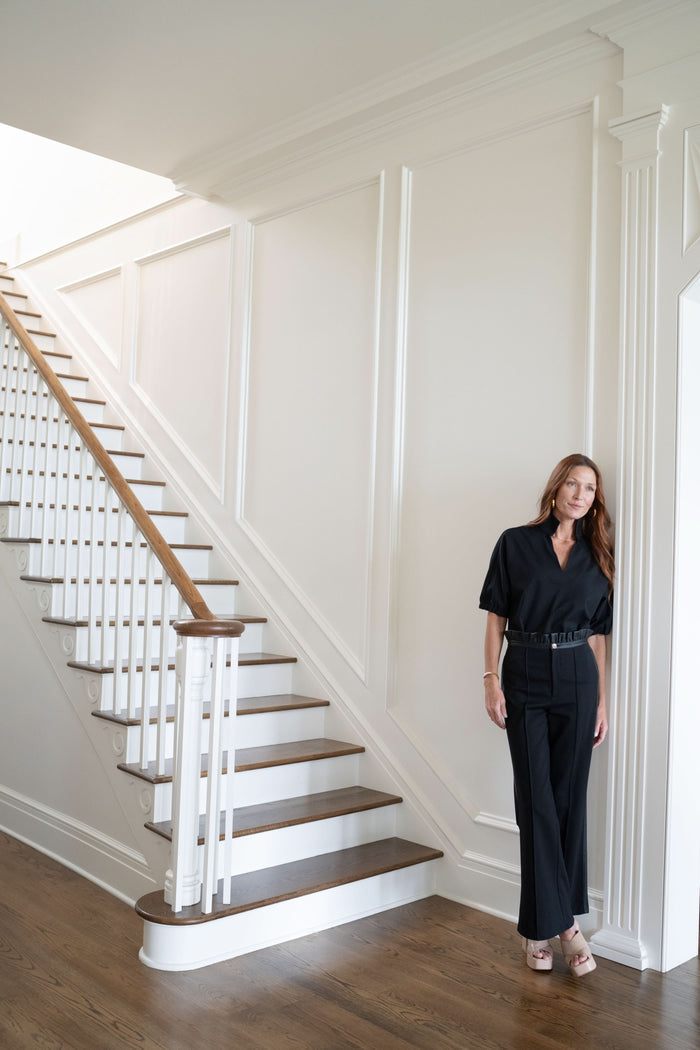A woman with long hair, wearing the Poppy Top and matching pants, stands leaning against a white wall at the foot of a wooden staircase in a modern, well-lit home.