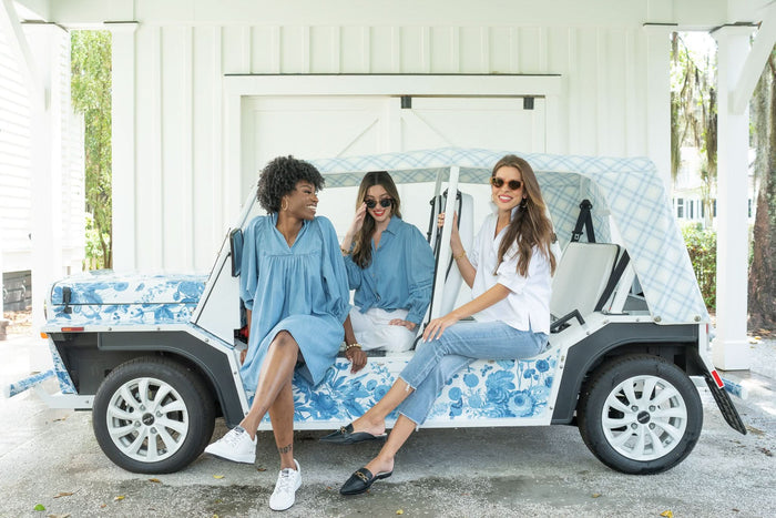 Three women smile under a white carport, sitting on a blue and white floral electric vehicle. All wear sunglasses and relaxed blue and white outfits, including the loose-fitting Poppy Top, with greenery visible in the background.