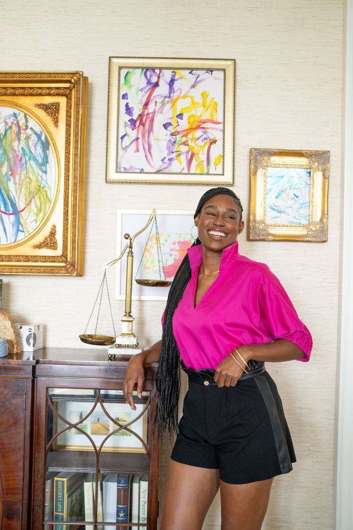 A person wearing a Poppy Top in Fuchsia Fedora Cotton Poplin and black shorts smiles while leaning against a wooden bookshelf. Behind them are colorful abstract paintings in gold frames on a beige wall, complemented by the decorative scale on the shelf.
