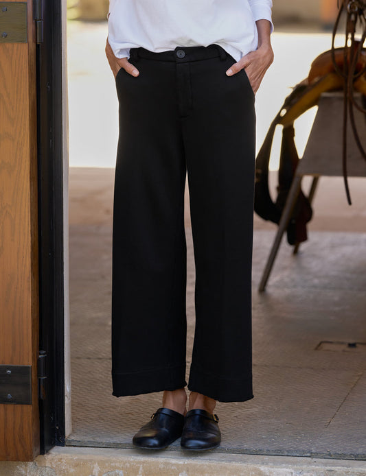 A person wearing black wide-leg cropped trousers with a white top and black loafers, standing in a doorway.