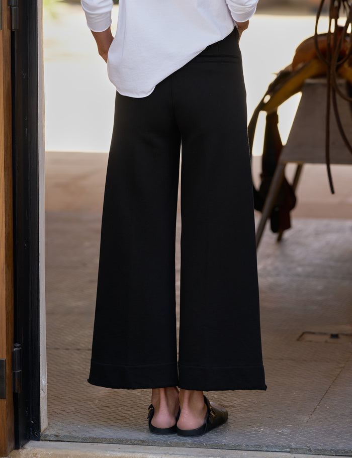 A person shown from the back wearing black wide-leg cropped pants with a white long-sleeve top and black sandals, standing in a doorway.