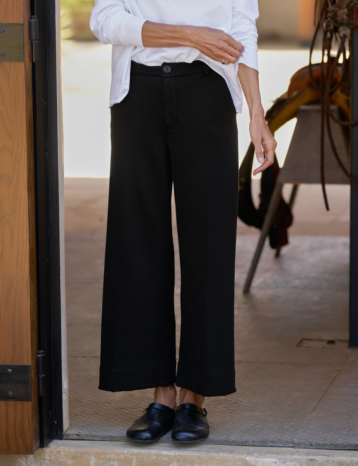 A person wearing black wide-leg cropped pants with a white button-up shirt and black loafers, standing in a doorway.