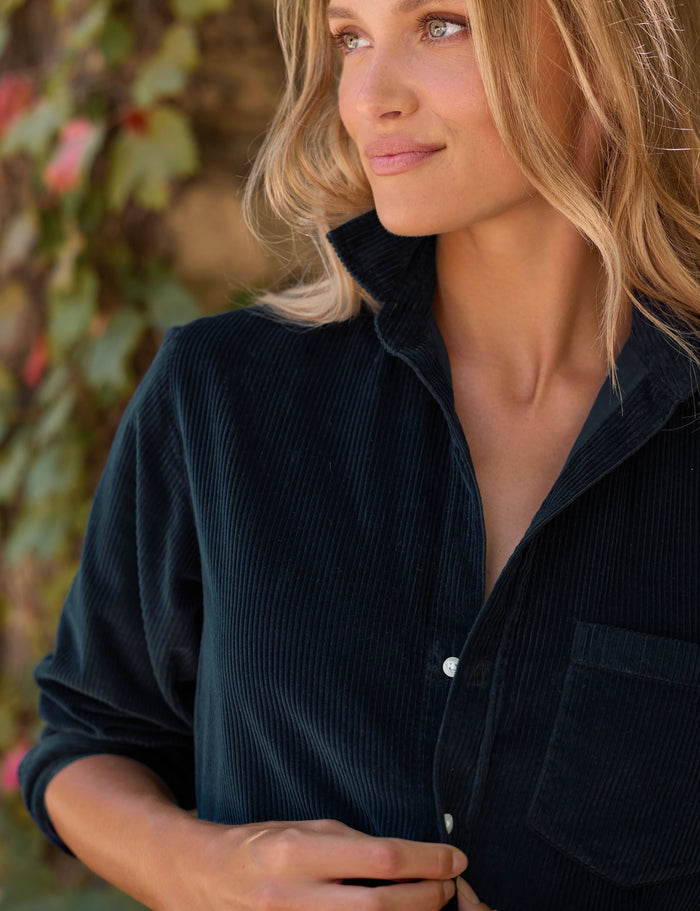 A close-up of a woman wearing a dark navy corduroy button-up dress, looking off-camera against a soft-focus background of green foliage.