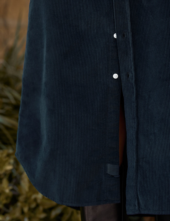 Close-up of a dark navy corduroy button-front garment, showing the ribbed texture, white buttons, and a side slit with a brown lining.