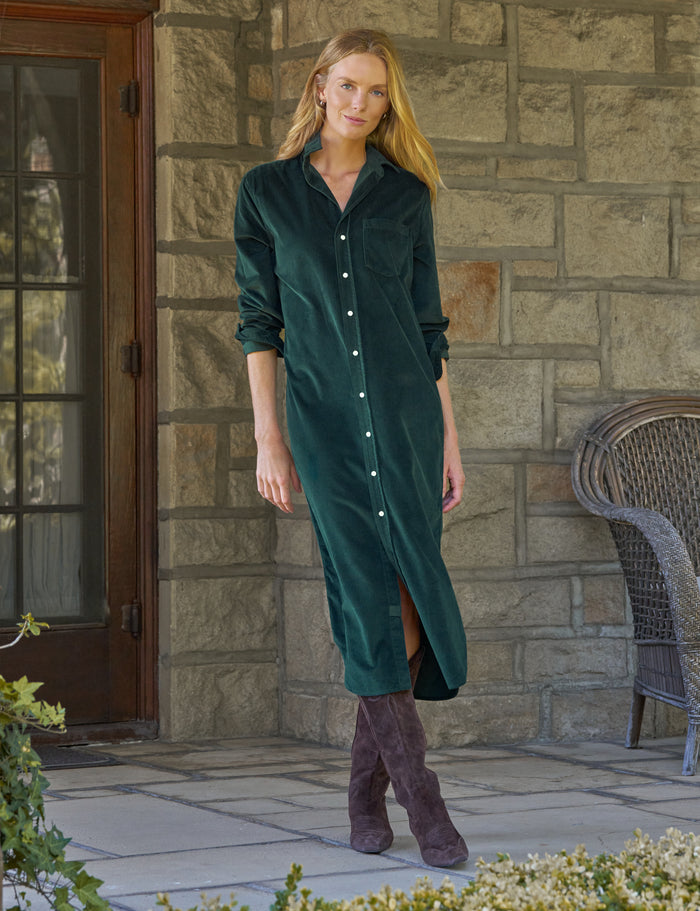 A woman stands on a stone patio wearing a dark green corduroy button-front maxi dress with long sleeves and brown suede boots. A stone wall, a glass door, and a wicker chair are visible in the background.
