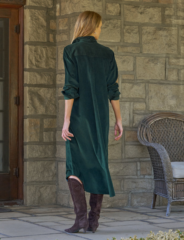 A woman stands with her back to the camera wearing a dark green corduroy maxi shirt dress and brown knee-high boots, posing in front of a stone wall with a wicker chair to the side.