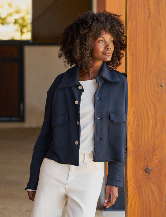 A woman with curly dark hair wearing a navy blue cropped utility jacket over a white top and white pants, standing next to a wooden pillar in an outdoor patio setting.