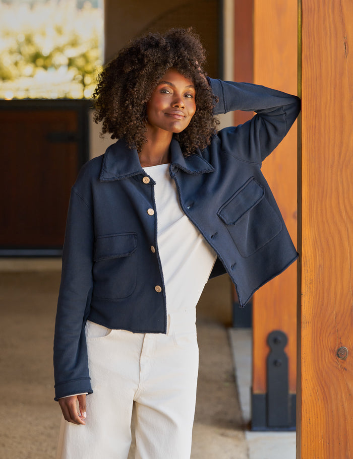 A woman with curly dark hair models a navy blue cropped jacket with gold buttons, worn over a white top and cream pants, leaning against a wooden door.