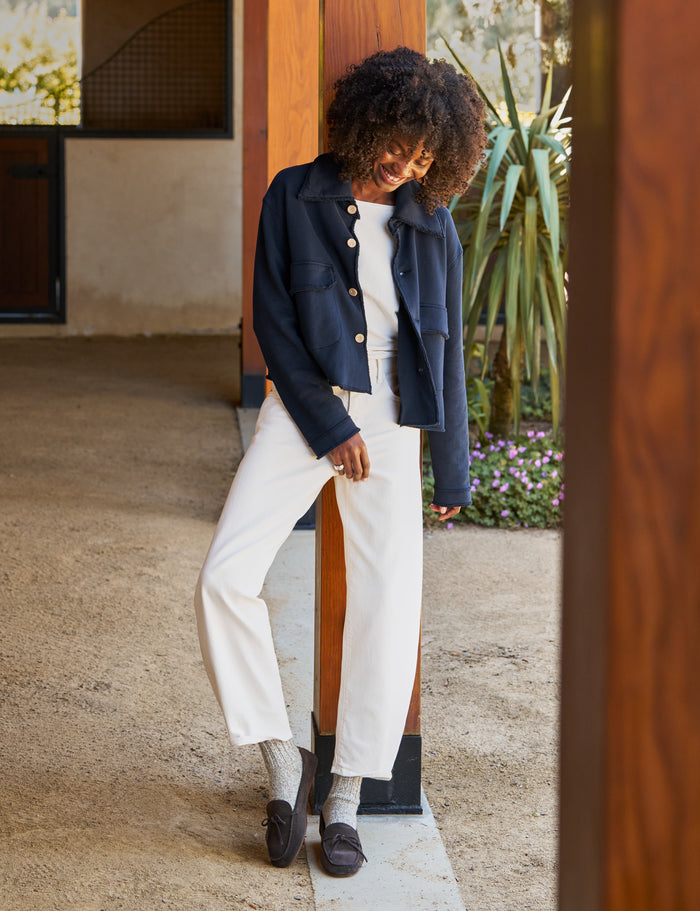 A woman wearing a navy blue cropped jacket, white top, and white wide-leg cropped pants, leaning against a wooden pillar in a modern outdoor corridor.