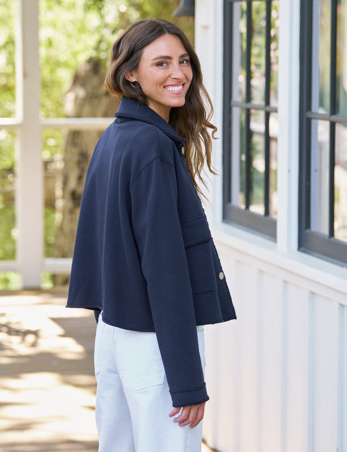 A woman wearing a navy blue cropped jacket and white pants, smiling and looking back over her shoulder in front of a white building with large windows.