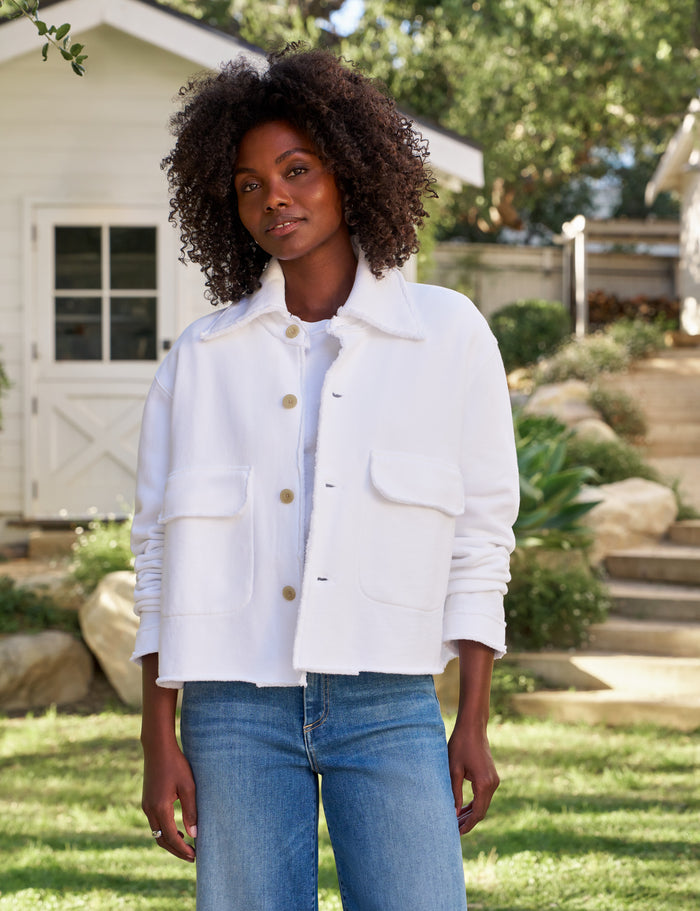 A woman wearing a white cropped jacket with front buttons and flap pockets, paired with blue jeans, standing outdoors in a garden setting.