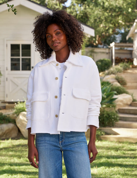 A woman wearing a white cropped jacket with front buttons and flap pockets, paired with blue jeans, standing outdoors in a garden setting.