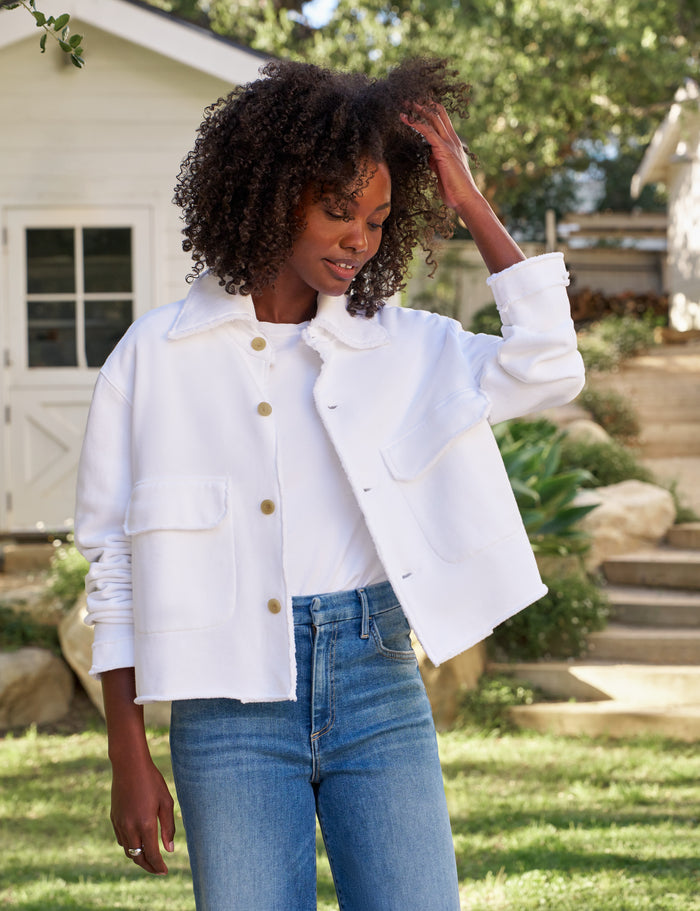 A woman wearing a white cropped jacket with gold buttons and blue jeans, standing outdoors in a garden setting.