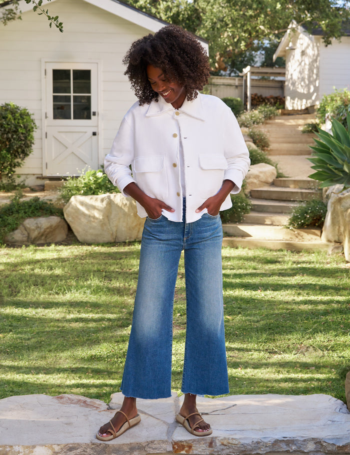 A woman stands outdoors in a grassy yard wearing a white cropped button-front jacket with front flap pockets, blue wide-leg cropped jeans, and brown sandals.