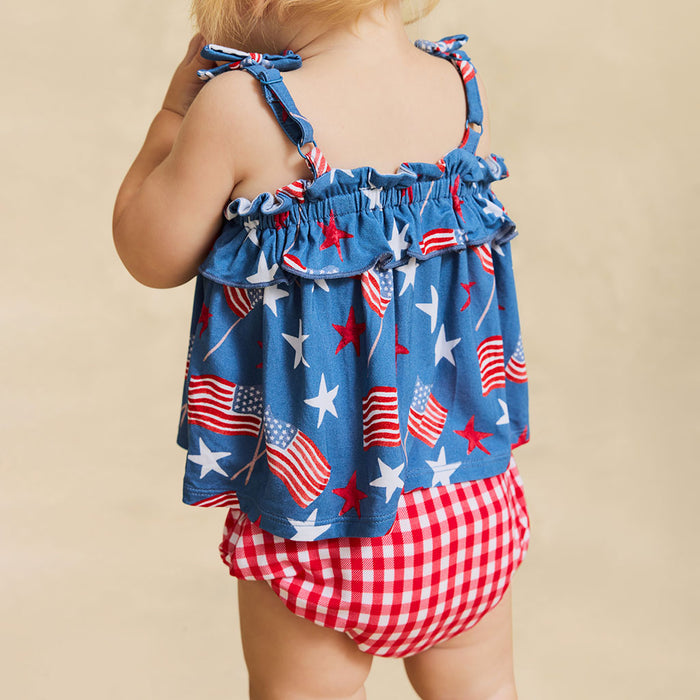 Child wearing a blue top with American flag pattern and red checkered shorts on a beige background