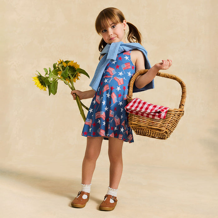 Young girl in a blue dress with American flag pattern, holding sunflowers and a picnic basket.