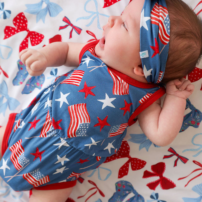Baby wearing a blue outfit with American flag pattern on a matching headband, lying on a colorful blanket.