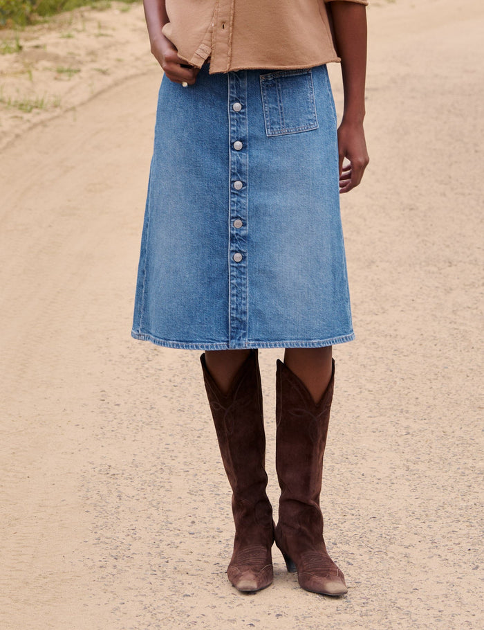 A woman stands on a dirt path in a rural outdoor setting, wearing a tan cropped button-front cardigan, a blue denim midi skirt, and brown knee-high boots.