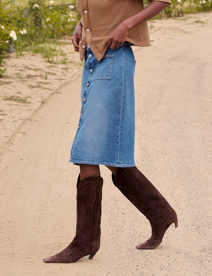 A woman walking on a dirt path outdoors, wearing a tan button-front cardigan, a light blue denim midi skirt, and dark brown heeled boots. A fence, green field, and trees are visible in the background.