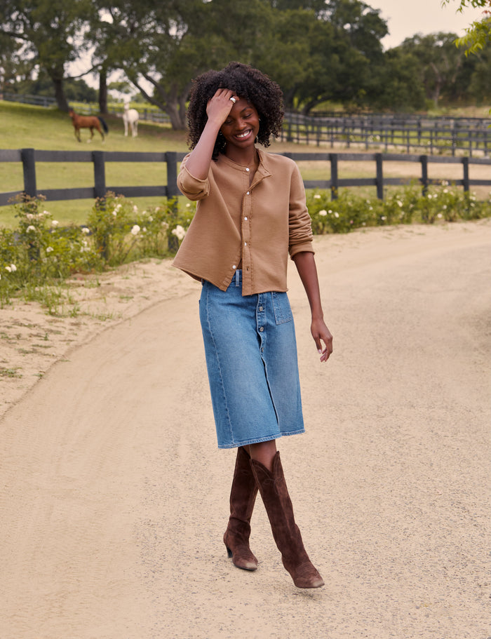 A woman stands on a dirt path outdoors wearing a tan cropped button-front cardigan, a blue denim midi skirt, and brown knee-high boots. A fence, green field, and trees are visible in the background.