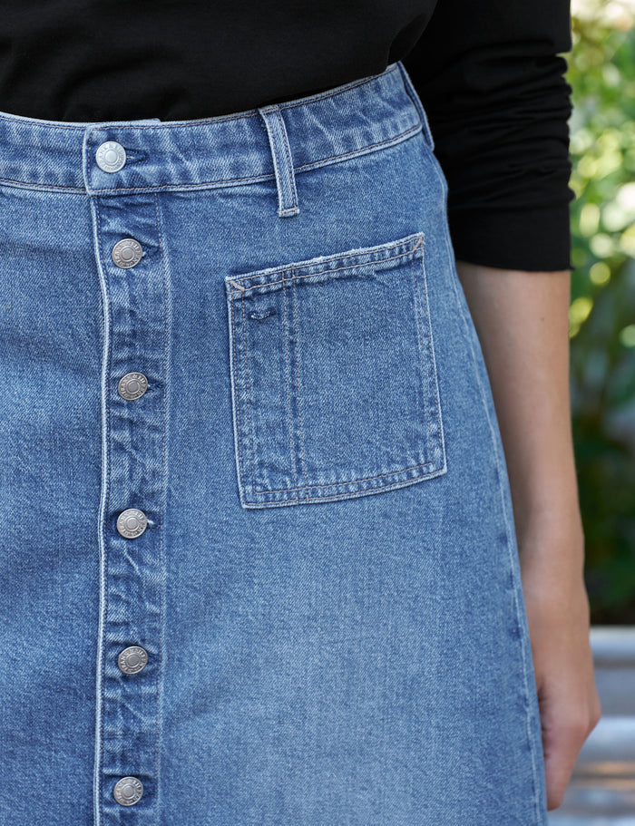 Close-up of a light blue denim skirt with a button-front closure and a front patch pocket, worn with a black long-sleeve top. A blurred outdoor background is visible on the right.