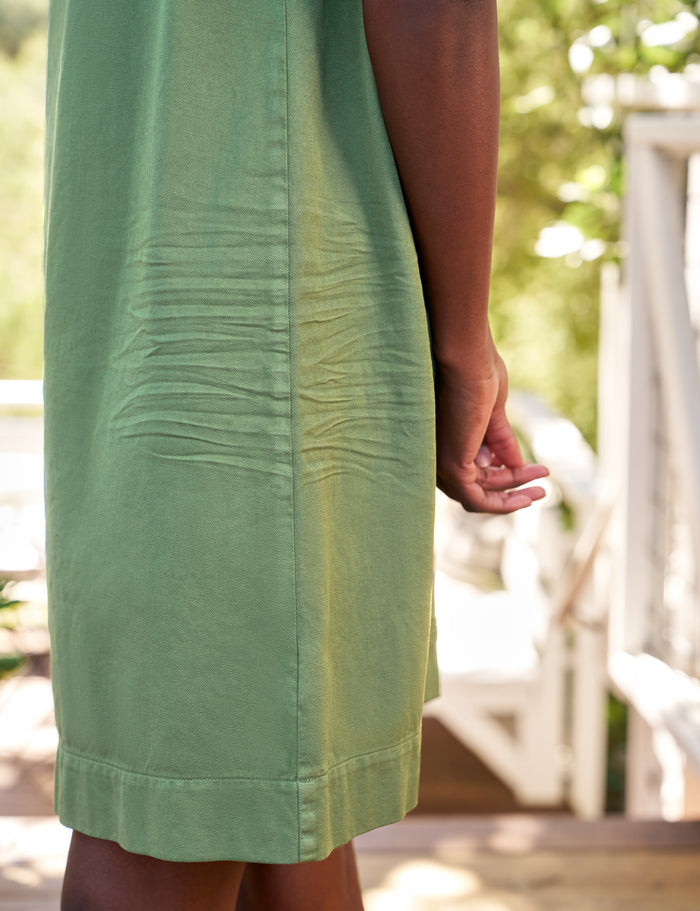 A close-up side view of a person wearing a light sage green dress outdoors, showing the side seam, hand, and part of the legs against a softly blurred porch background.
