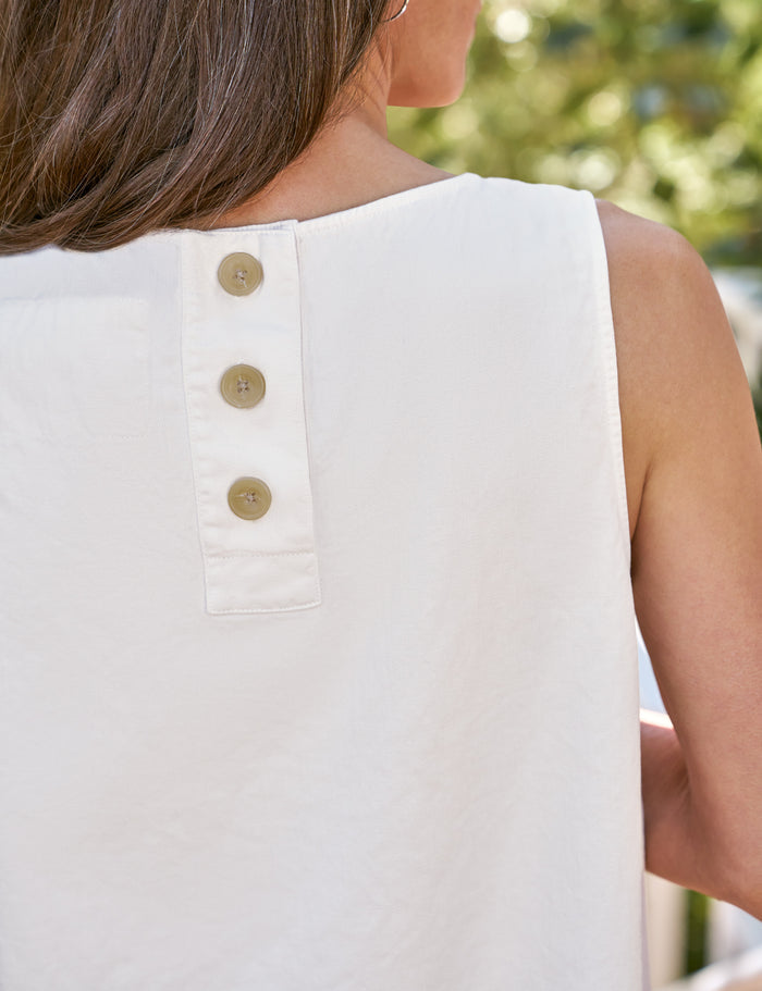 Close-up back view of a woman wearing a sleeveless white top with three gold buttons down the center back, set against a blurred outdoor background.