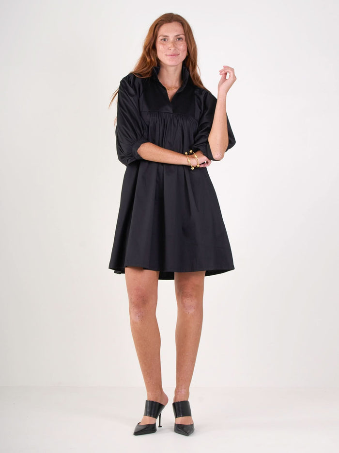 A woman with long, wavy reddish-brown hair wears the Stella Dress from Emily McCarthy Collection with three-quarter sleeves and black pointed heels, standing against a plain white background.