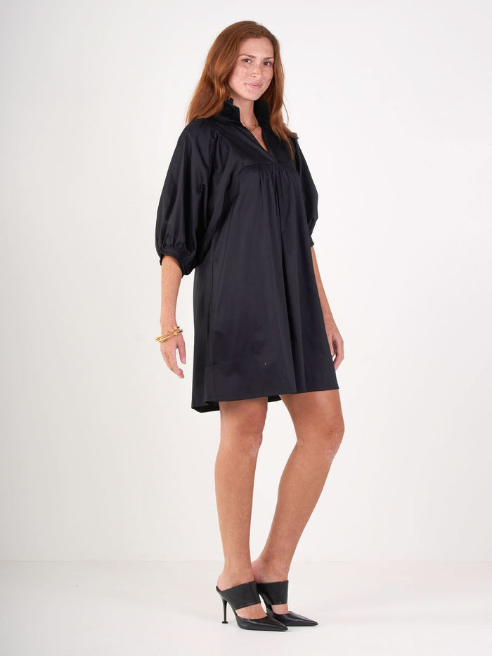 A woman with long red hair wears the Stella Dress—a loose-fitting black dress with elbow-length puff sleeves—from Emily McCarthy Collection, paired with black heels, standing against a plain white background.