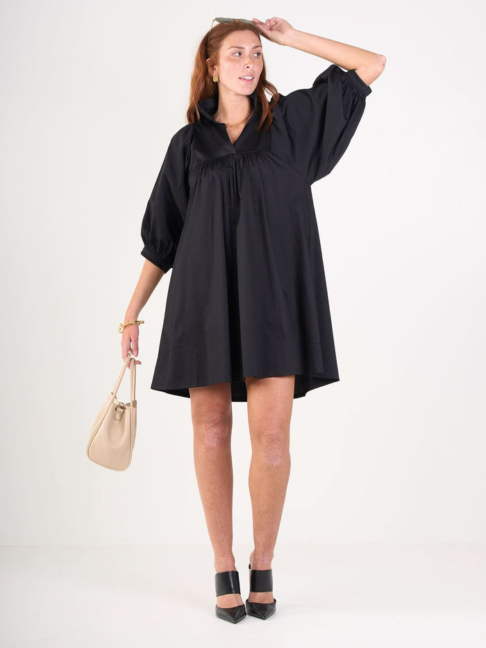 A woman wears the Stella Dress from Emily McCarthy, made of black cotton poplin with three-quarter sleeves, posing against a white background. She holds a beige handbag, wears black heels, and has auburn hair styled in a ponytail.