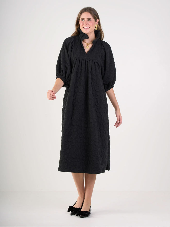 A woman smiles against a plain background, wearing the loose-fitting Stella Midi dress with embossed cheetah texture and elbow-length puff sleeves, paired with black bow flats plus gold earrings and necklace.