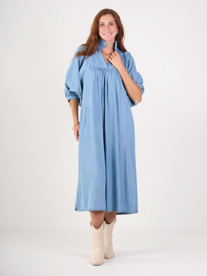 A person with long red hair smiles in a Stella Midi dress with a ruffle stand collar and light blue puffy sleeves paired with stylish white cowboy boots, against a plain white background.