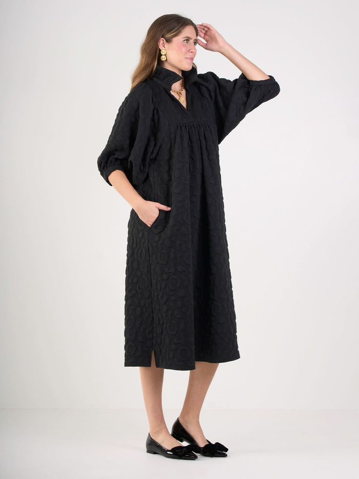 A woman stands sideways in the Stella Midi, a black embossed cheetah print, knee-length dress with three-quarter sleeves, pockets, and a ruffled stand collar. She pairs it with black flats and gold earrings, one hand in her pocket, the other touching her hair.