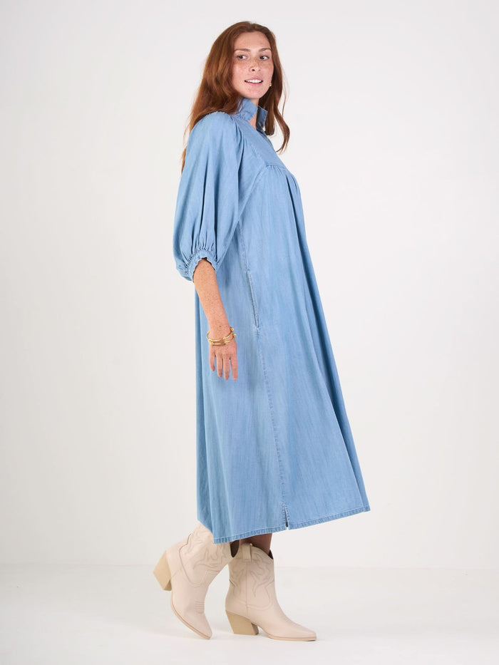 A person with long hair models the Stella Midi, a light blue, long-sleeved denim chambray dress with a ruffle stand collar. Paired with beige cowboy boots, they pose against a plain white background, turned slightly to the side with one foot forward.